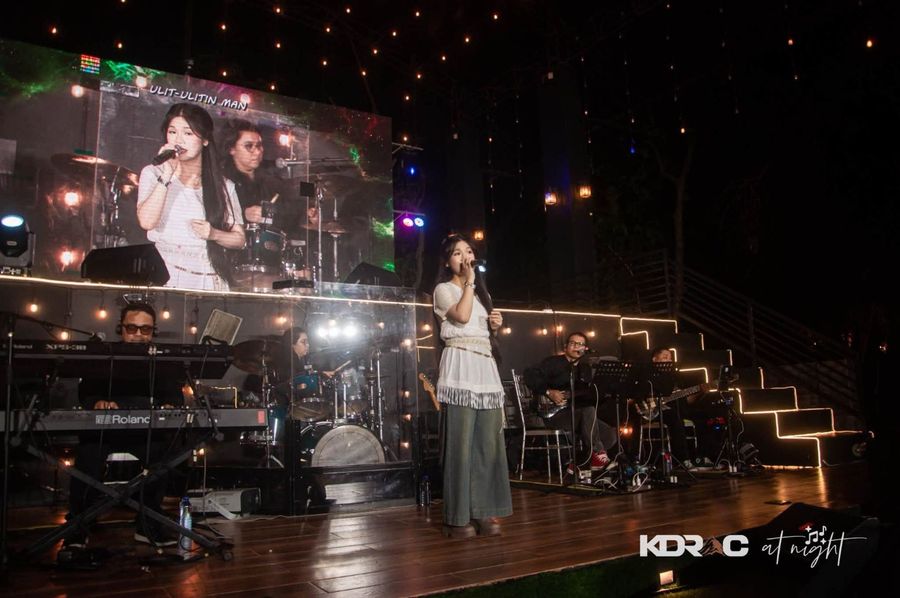 Young singer on the Pavilion stage with her band, the big screen and string lights behind
