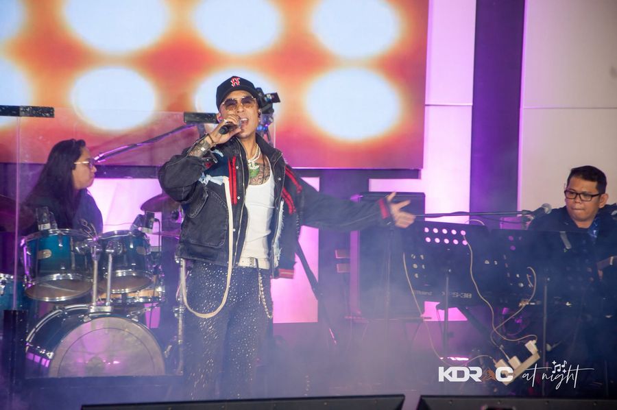 Performer in a bold jacket on stage under the lights, band behind